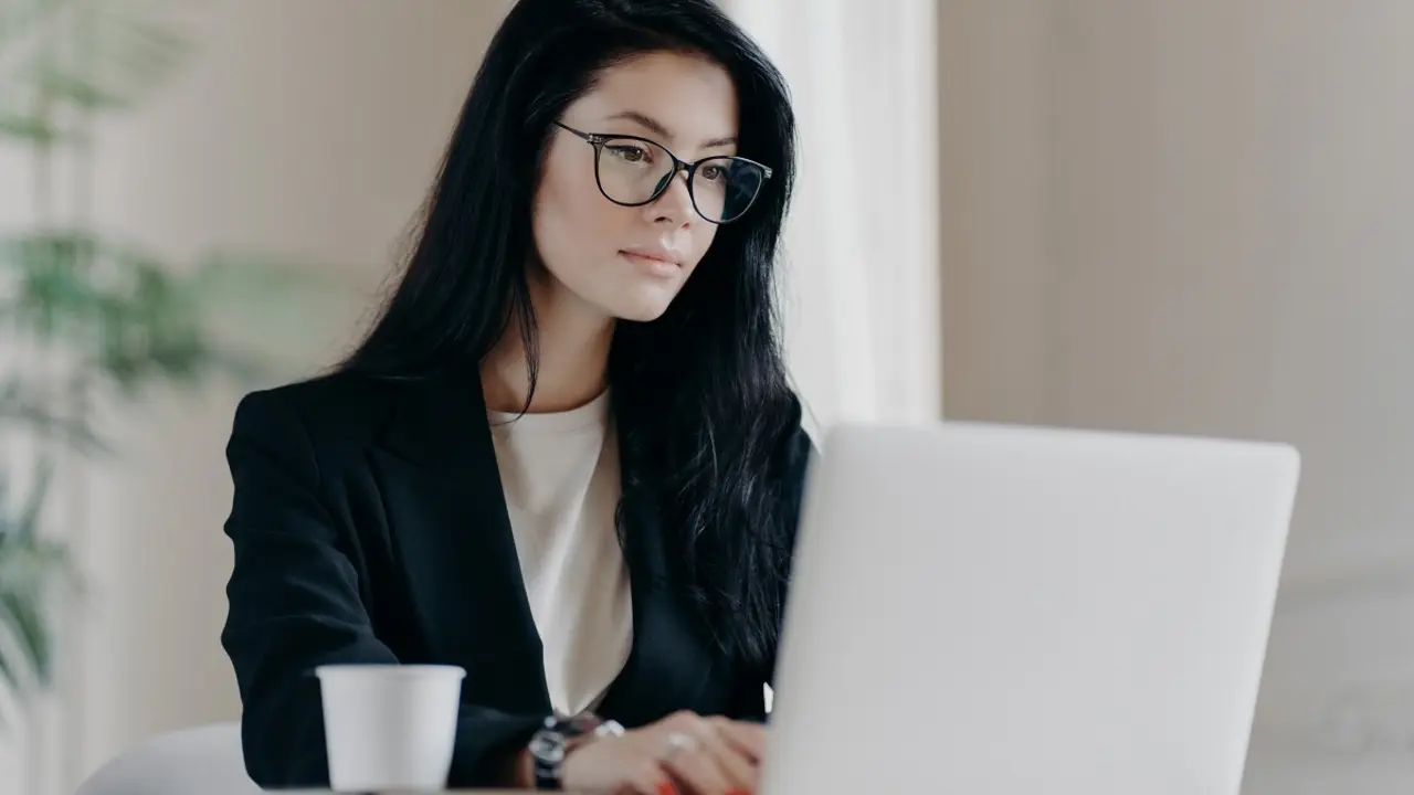 Concentrated brunette woman IT specialist in eyewear searches information using internet, keyboards on laptop computer, drinks coffee, has serious expression. Business and freelance concept.