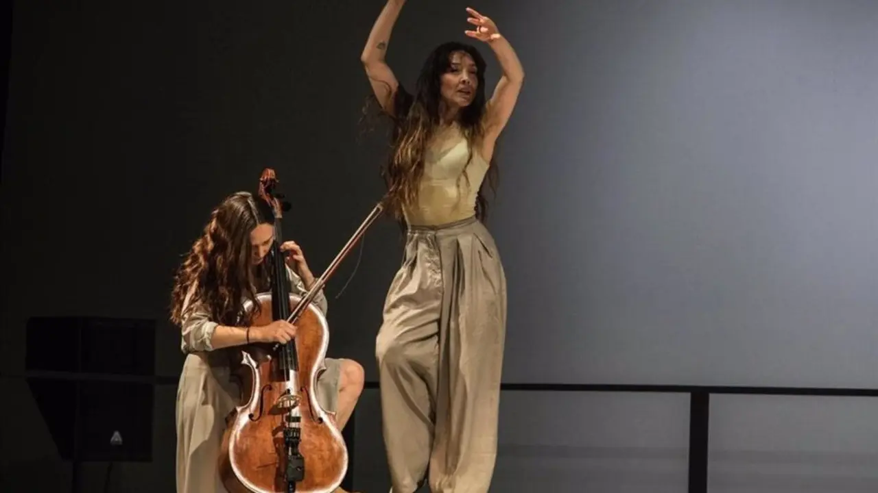 Escena de la obra Ant&iacute;podas donde puede apreciarse una mujer bailando sobre una mesa, o un espacio alto, y a su lado otra mujer tocando un violoncello